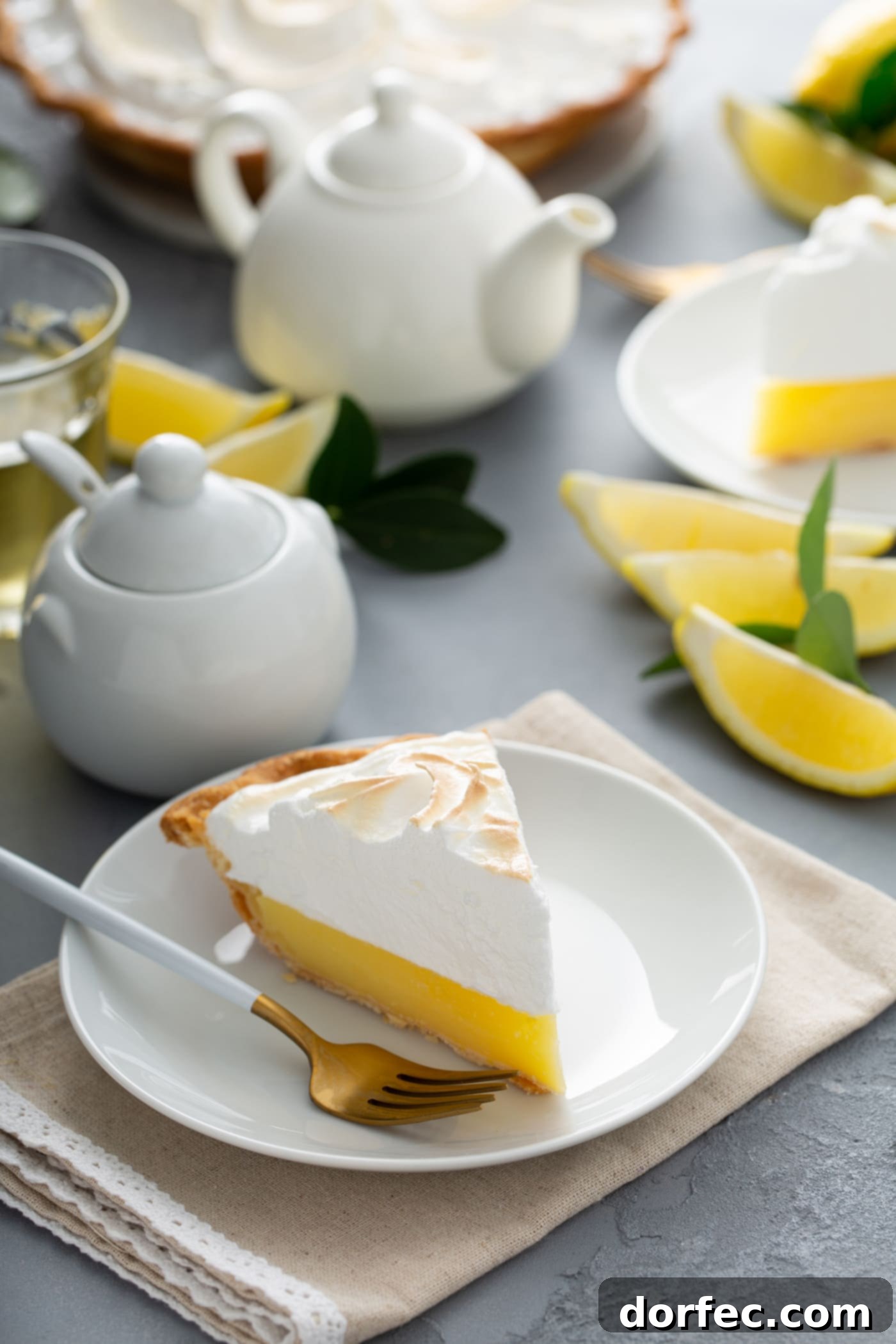 Mom's Lemon Meringue Pie A piece of lemon meringue pie on a dessert plate. A fork in next to the pie and there are lemon wedges and a sugar bowl and cream pitcher in the background.