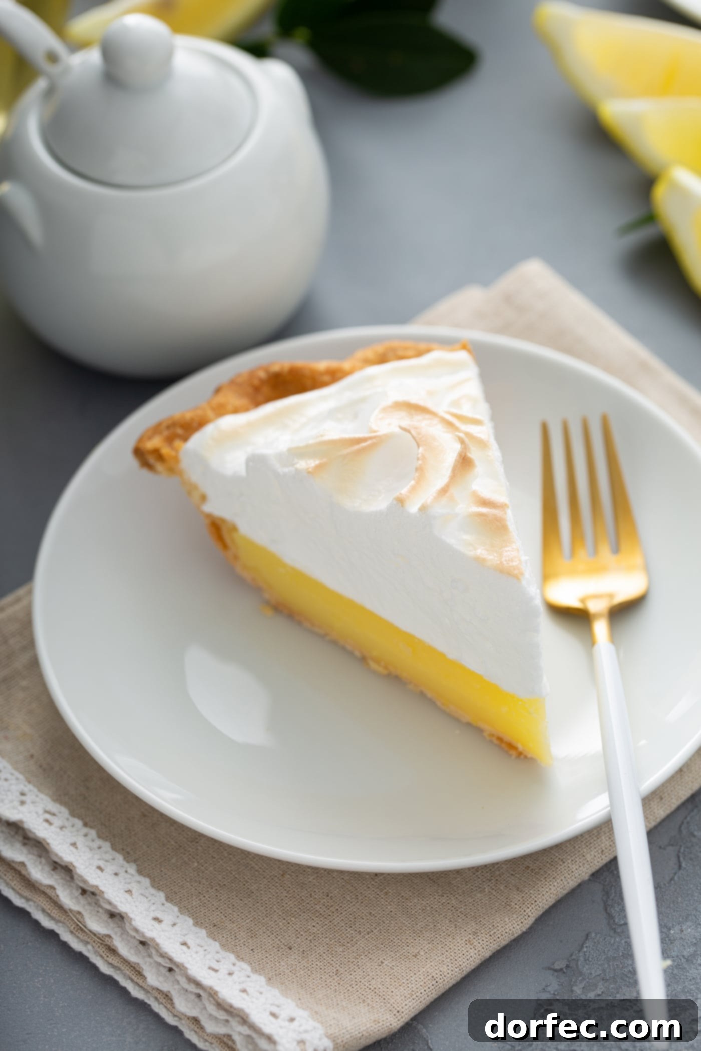 Mom's Lemon Meringue Pie A slice of lemon meringue pie on a white dessert plate with a fork next to it. A sugar bowl is in the background.