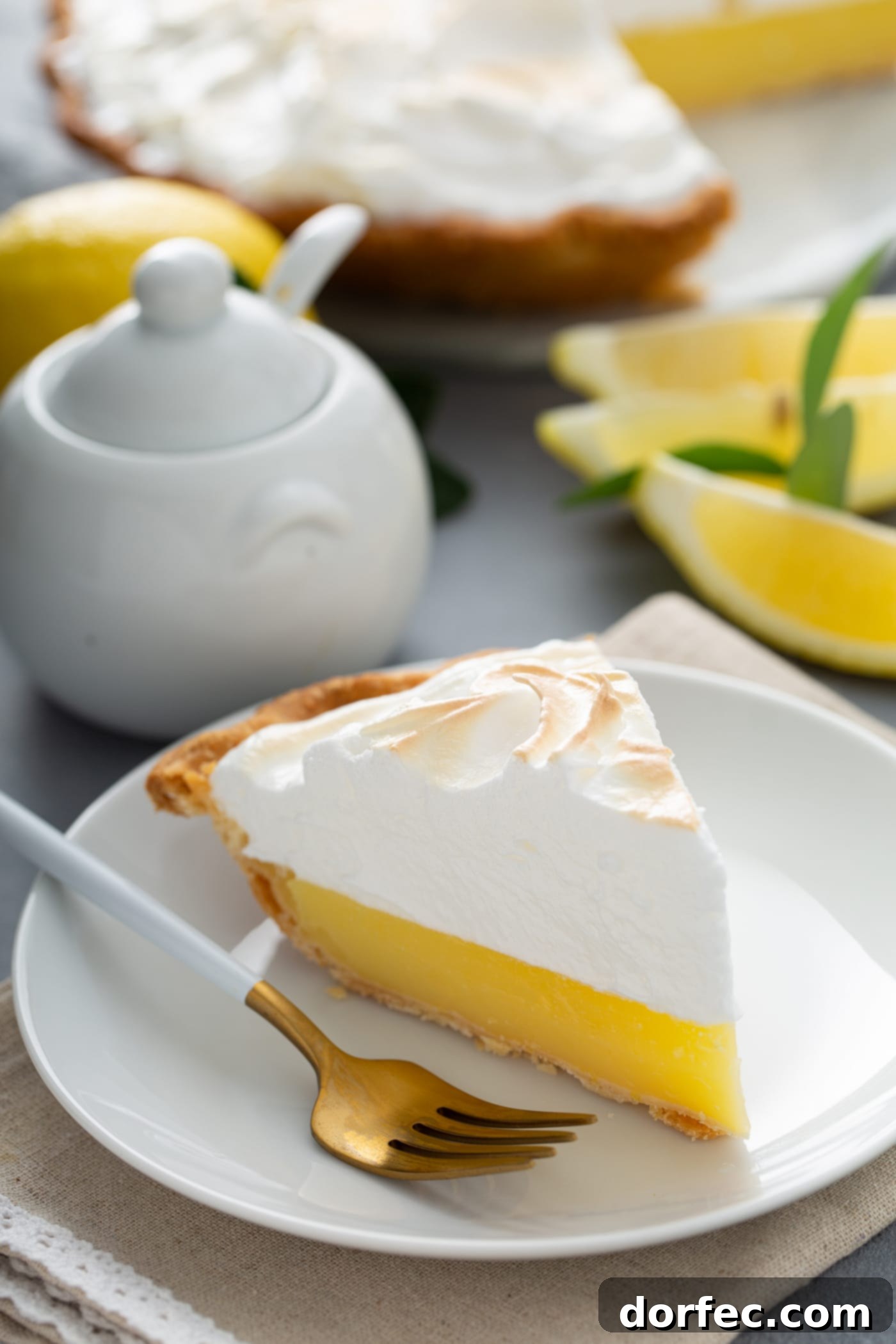Mom's Lemon Meringue Pie A picture of a slice of lemon meringue pie. The filling is bright yellow and the meringue on top is lightly toasted. There are lemon wedges and a sugar bowl in the background and a fork is on the plate next to the slice of pie.