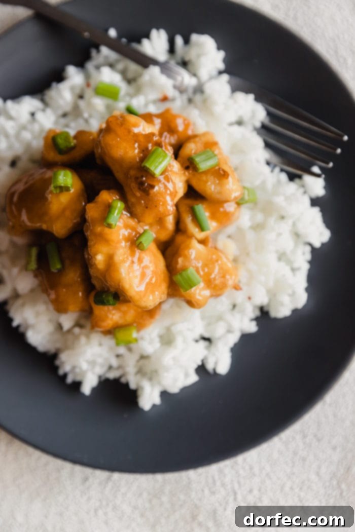A dark plate with fluffy white rice and orange chicken sprinkled with green onions over the top and a white bowl with extra orange sauce in the background