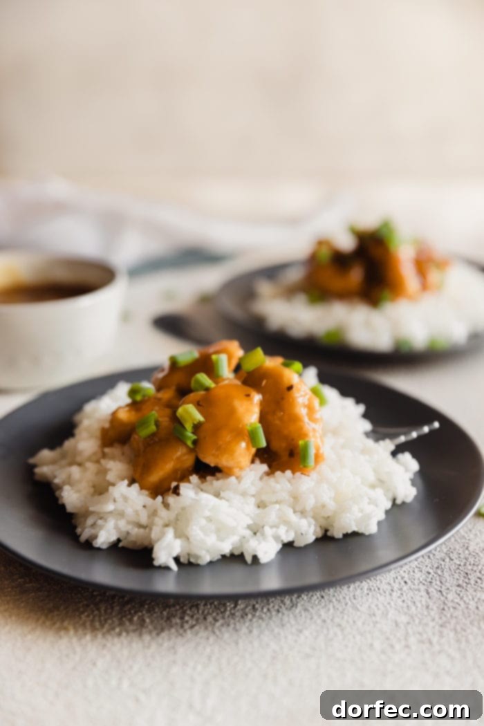 A dark plate with fluffy white rice and orange chicken sprinkled with green onions over the top and a white bowl with extra orange sauce in the background