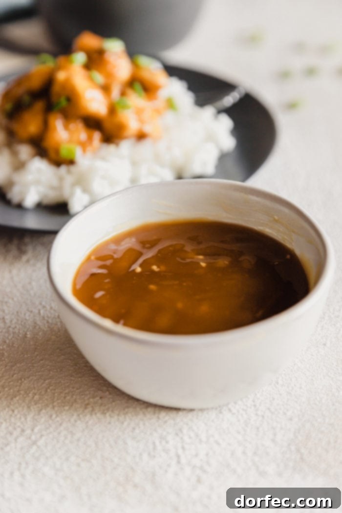 a white bowl with easy asian orange sauce for chicken and a grey plate with white long grain rice topped with orange chicken and green onions in the background