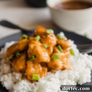 A dark plate with fluffy white rice and orange chicken sprinkled with green onions over the top and a white bowl with extra orange sauce in the background