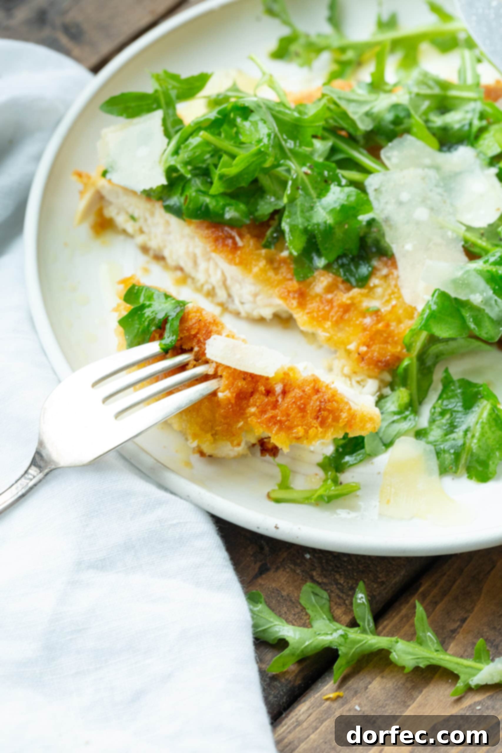 A close-up photo of a thinly sliced, golden breaded chicken breast, with a fork removing a bite. The chicken is adorned with vibrant salad greens and delicate shavings of Parmesan, highlighting the fresh and light nature of the dish.