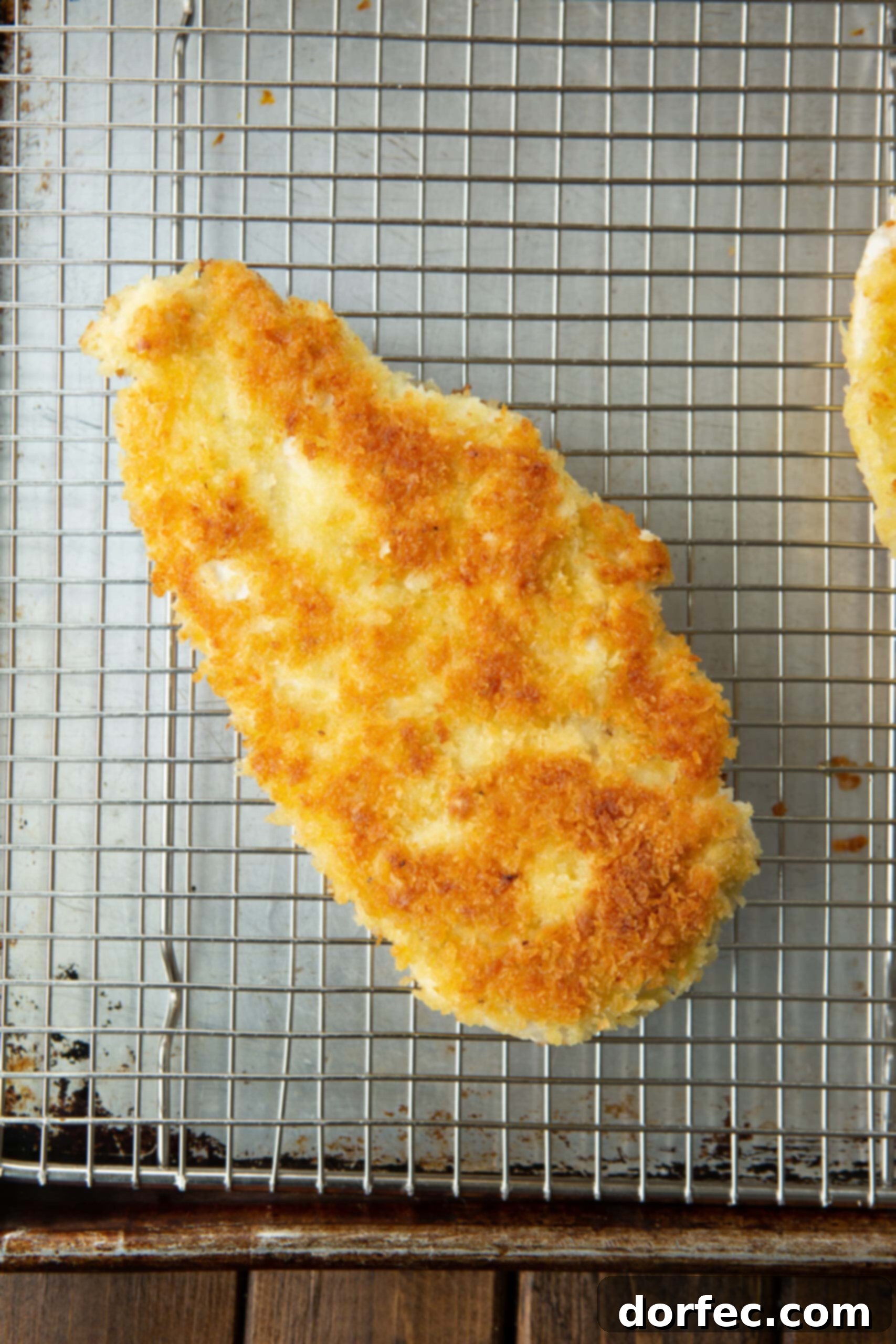 A close-up photograph of a single pan-seared, golden breaded chicken breast, elegantly resting on a wire cooling rack, ready for serving. Its perfectly crisp crust hints at the deliciousness within.