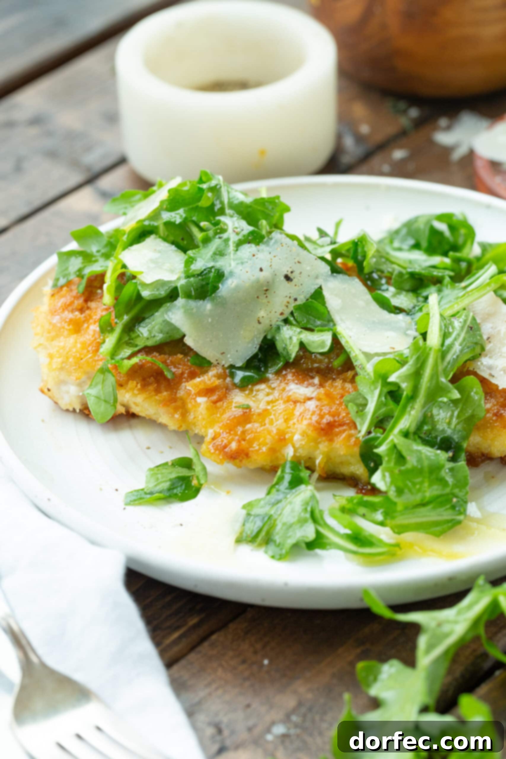 A golden lightly breaded chicken breast artfully arranged on a white dinner plate, garnished with a medley of fresh salad greens and delicate shavings of Parmesan cheese.