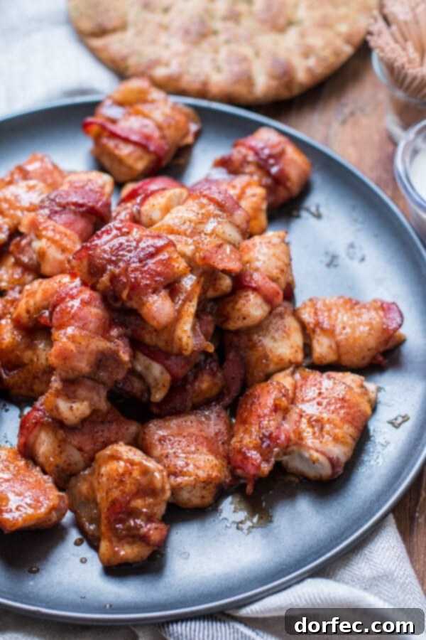 Serving of Baked Bacon-Wrapped Chicken Bites pile of crispy bacon wrapped chicken breast pieces on a gray plate, garnished with fresh herbs
