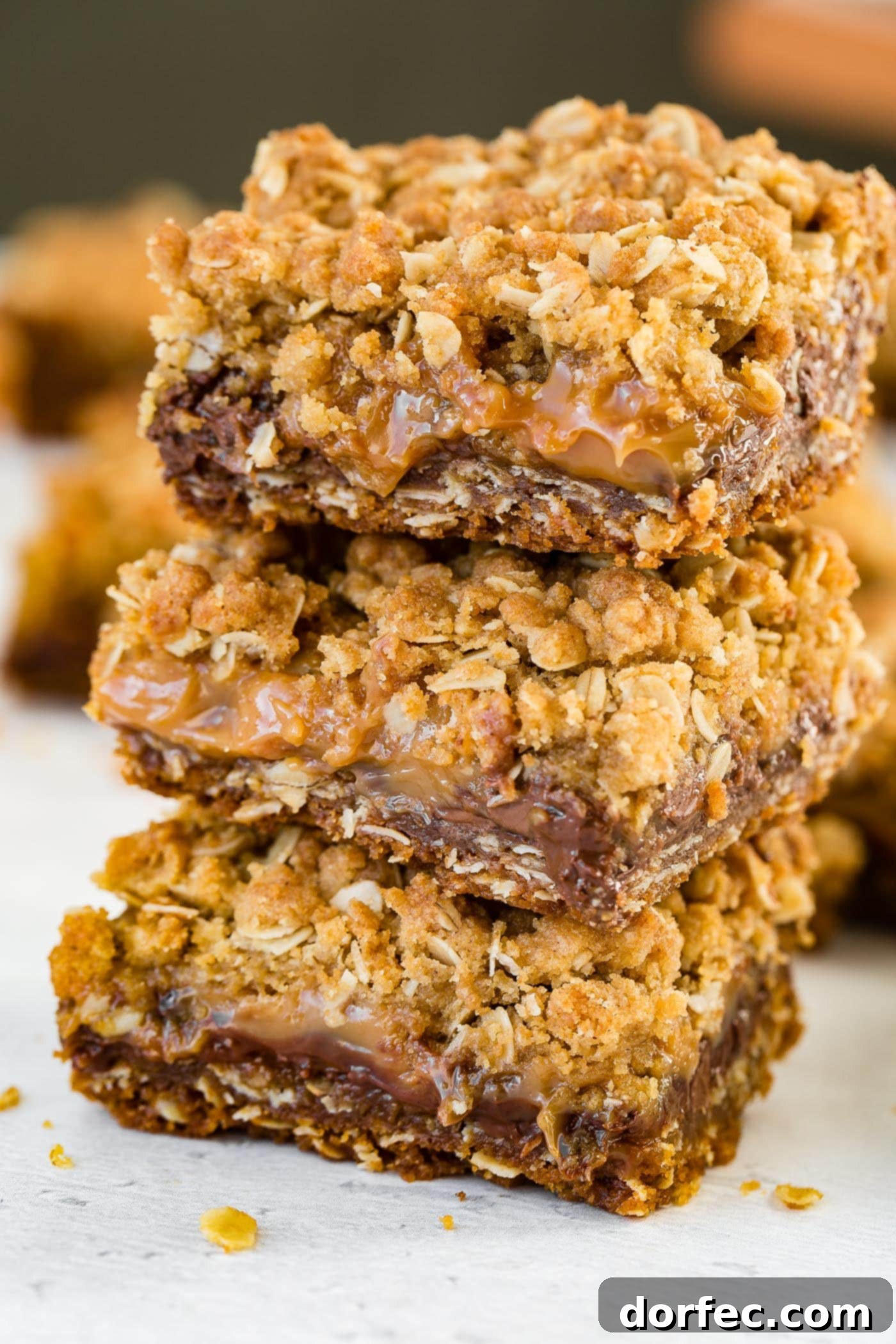 A photo of three oatmeal Carmelita bars stacked on a plate, with gooey caramel and silky chocolate visible, against a warm, inviting background.