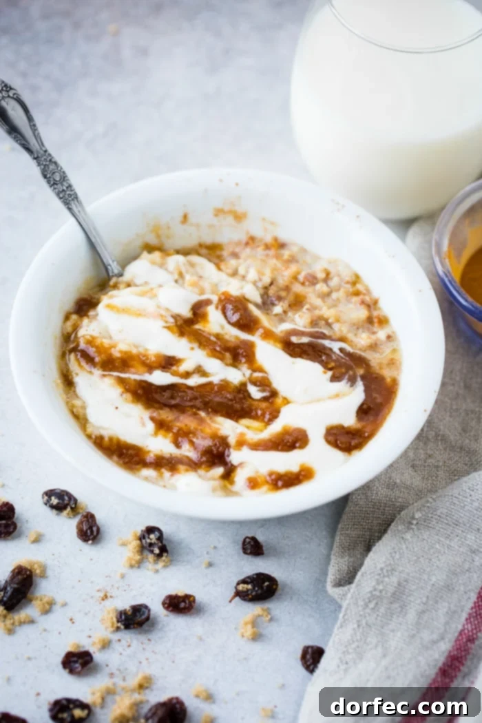 A bowl of lightened up cinnamon roll oatmeal, garnished with a swirl of cinnamon filling and a creamy glaze, surrounded by sprinkles of brown sugar, cinnamon, and raisins.
