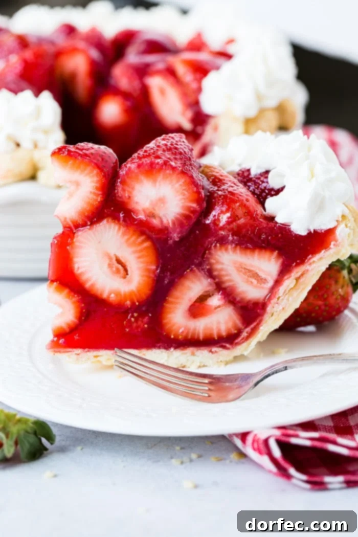 Irresistible Fresh Strawberry Pie 2 A white plate with a big slice of fresh strawberry pie made with jello and a little stabilized whipped cream with a red and white checkered napkin underneath and the whole pie in the background