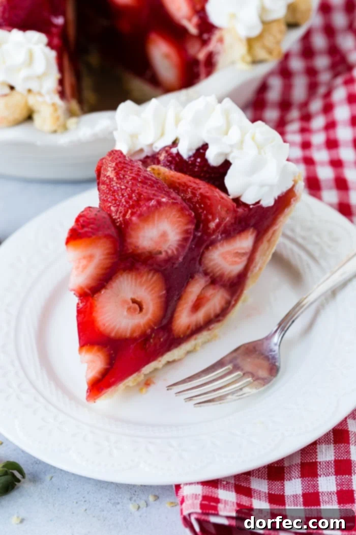 Irresistible Fresh Strawberry Pie 3 A white plate with a big slice of fresh strawberry pie made with jello and a little stabilized whipped cream with a red and white checkered napkin underneath and the whole pie in the background