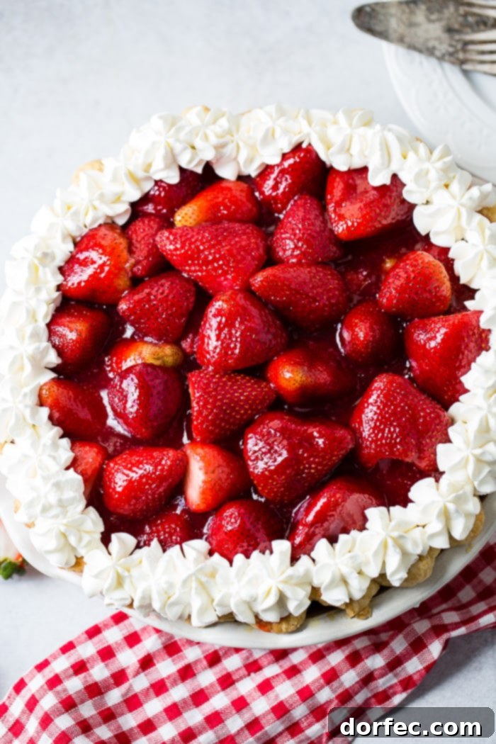 Irresistible Fresh Strawberry Pie 5 a white background with a red and white checkered napkin sitting under a whole fresh strawberry pie with stabilized whipped cream piped around the edges
