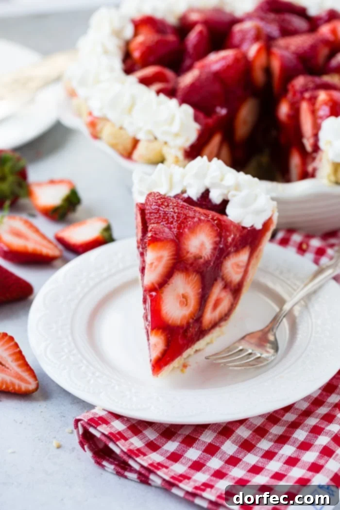 Irresistible Fresh Strawberry Pie 7 A white plate with a big slice of fresh strawberry pie made with jello and a little stabilized whipped cream with a red and white checkered napkin underneath and the whole pie in the background