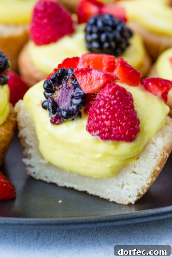 Sunshine Lemon Sugar Cookie Bites 3 A dark pan showcasing multiple sugar cookie cups, each generously filled with a creamy lemon mixture and adorned with an assortment of blackberries, raspberries, and chopped strawberries for a vibrant summer dessert.