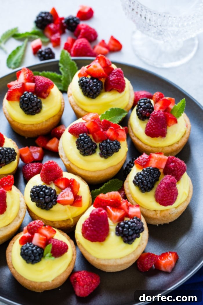 Sunshine Lemon Sugar Cookie Bites 4 A dark pan showcasing a row of six sugar cookie cups, each filled with a creamy lemon pudding mixture and generously topped with a colorful array of blackberries, raspberries, and chopped strawberries.