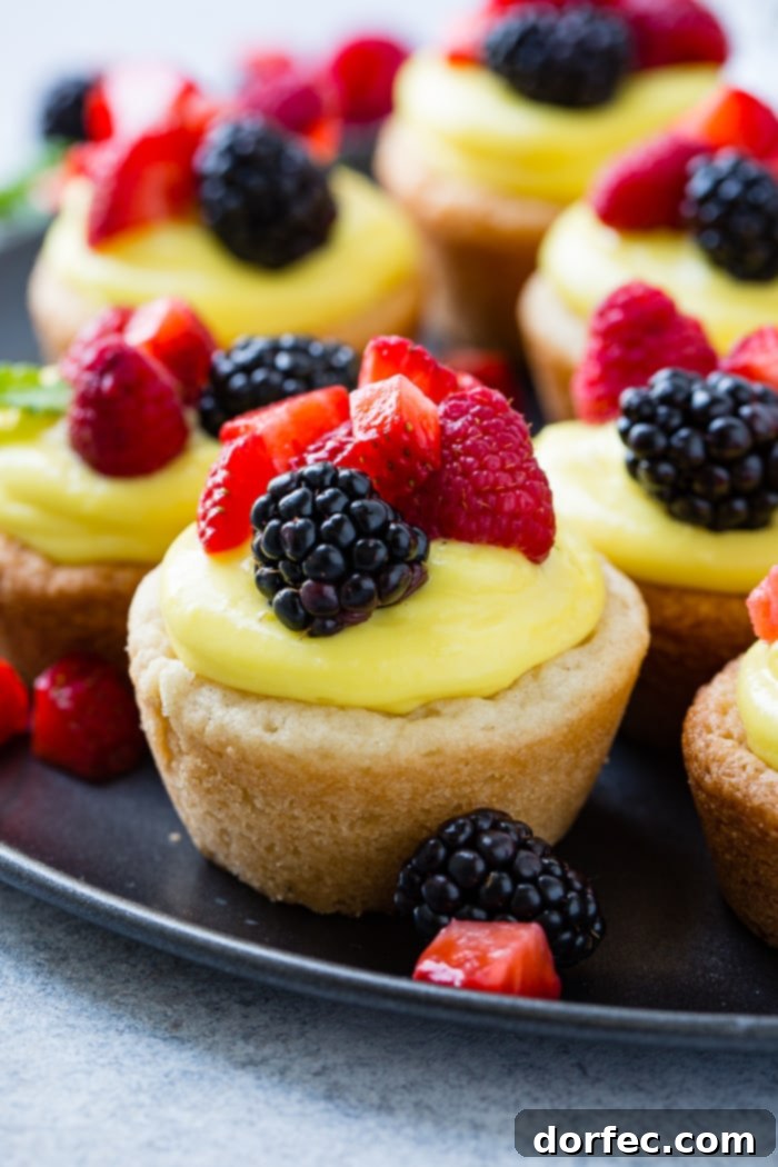 Sunshine Lemon Sugar Cookie Bites 5 A dark pan with a close-up view of a single sugar cookie cup, filled with a creamy lemon mixture and topped with blackberries, raspberries, and chopped strawberries, highlighting its vibrant and inviting appearance.