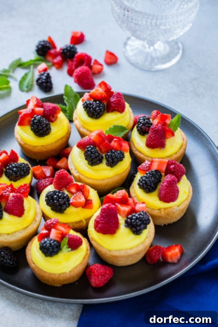 Sunshine Lemon Sugar Cookie Bites 7 A dark pan with a single sugar cookie cup, exquisitely detailed with a creamy lemon filling and topped with a vibrant assortment of blackberries, raspberries, and chopped strawberries, ready for a delicious bite.
