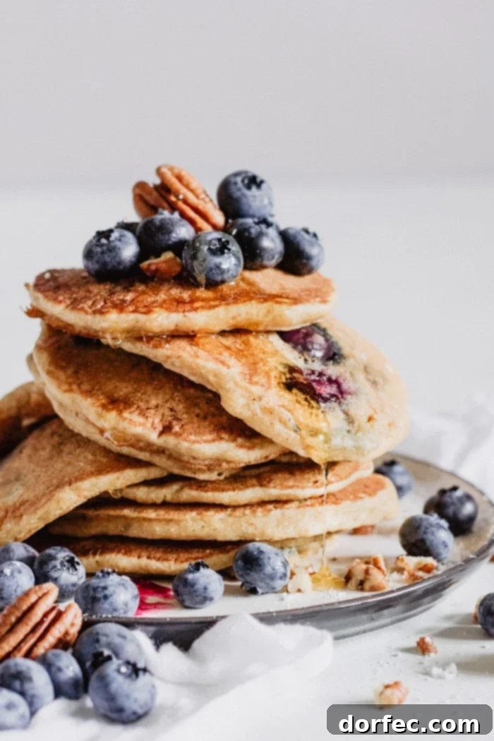 A beautifully arranged stack of golden brown blueberry pecan pancakes, generously drizzled with rich, amber maple syrup, invitingly placed on a white plate ready for a gourmet breakfast experience.