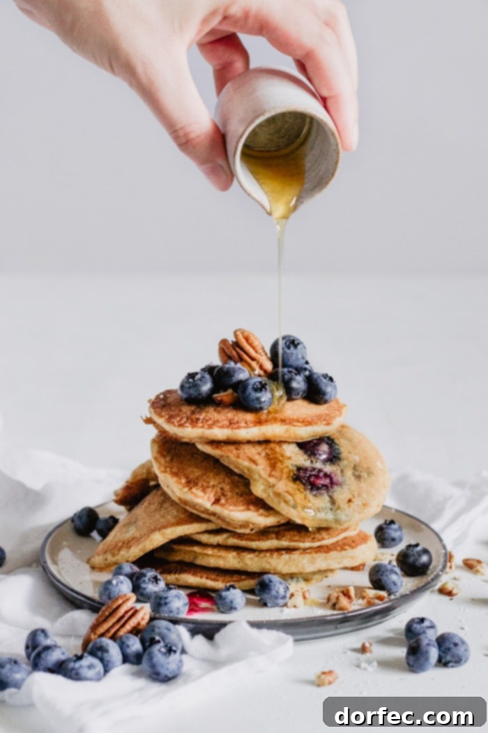 A visually appealing stack of golden brown blueberry pecan pancakes, beautifully presented on a rustic wooden board, lavishly adorned with extra fresh blueberries, chopped pecans, and a cascading stream of rich maple syrup.