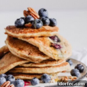 A vibrant, close-up image of a stack of freshly cooked blueberry pecan pancakes, adorned with extra blueberries and chopped pecans, and a generous pour of maple syrup, highlighting the textures and rich colors.