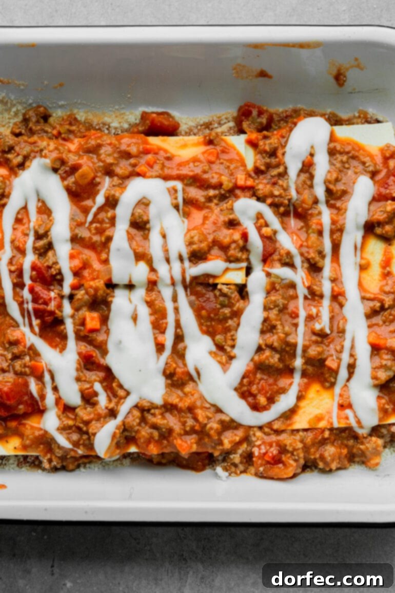 a photo of a lasagna being assembled in a baking dish with layers of noodles, meat sauce and a creamy bechamel sauce drizzled on top