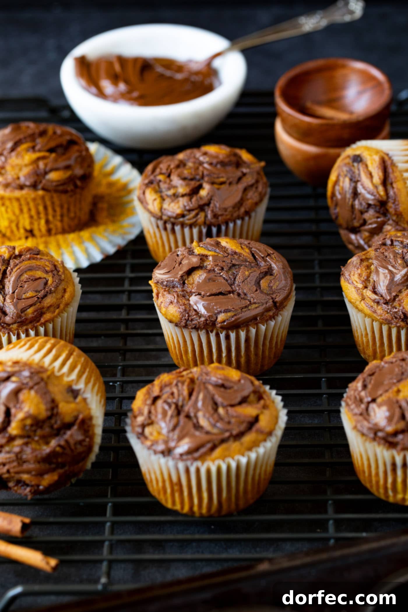 Nutella Swirled Pumpkin Muffins - Fall Breakfast Treat Ten Nutella swirled pumpkin muffins are artfully arranged. The paper liner has been gently peeled back on one of the muffins, revealing its soft, pumpkin-infused interior marbled with rich Nutella. A small bowl of creamy Nutella rests in the background, hinting at the key ingredient.