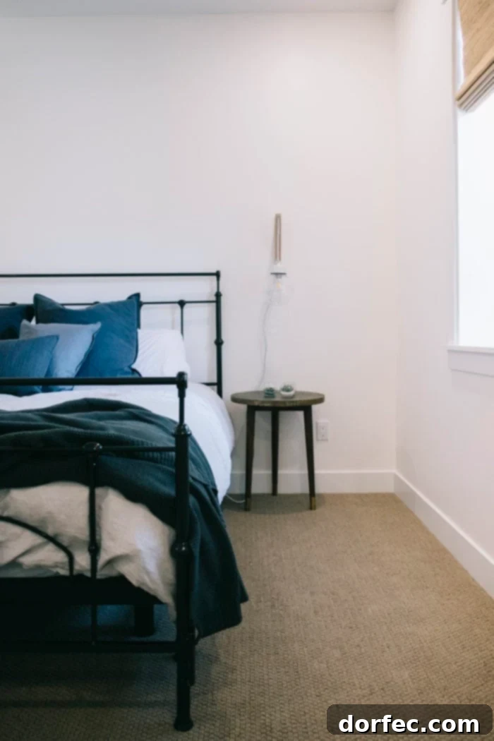 A chic basement bedroom featuring a black iron bed frame, light grey duvet, and an assortment of grey, blue, and white pillows. A minimalist nightstand holds a glass globe light hanging elegantly above it, creating a serene ambiance.