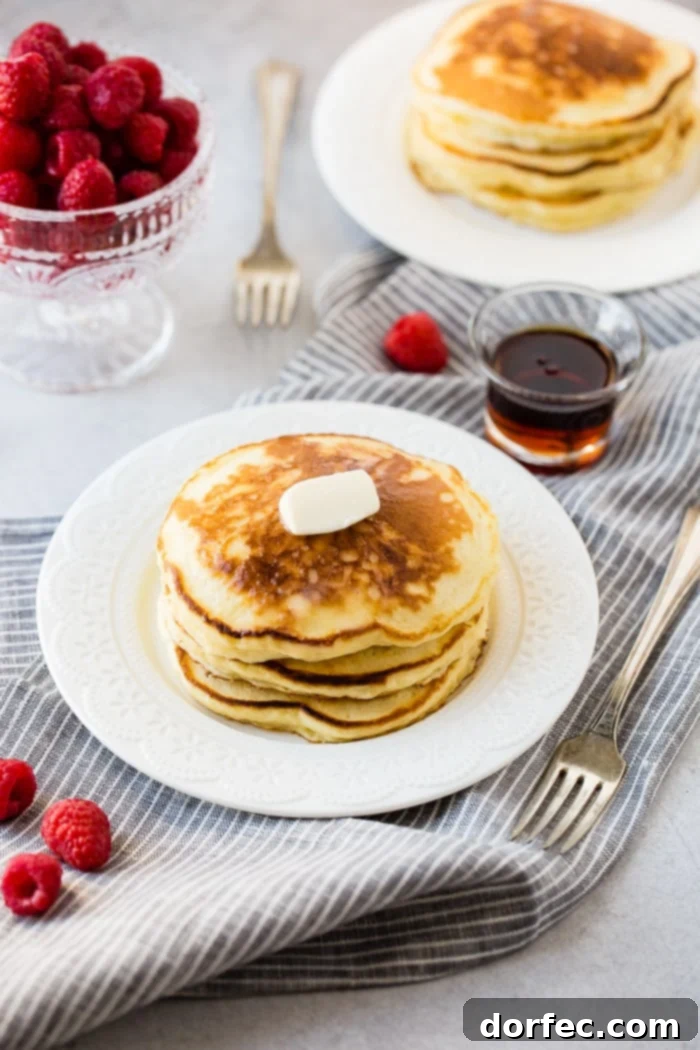 A stack of fluffy banana sour cream pancakes with a little butter melting on top and a drizzle of syrup dripping down the side. Our recipe for melt in your mouth sour cream pancakes on a striped napkin with raspberries and maple syrup.