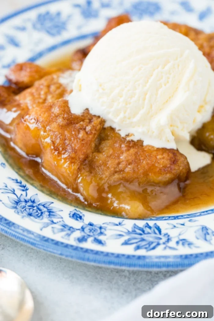 Sweet Simple Sprite Apple Dumplings 2 Two golden-brown apple dumplings, drizzled with caramel sauce and topped with a scoop of melting vanilla ice cream, presented on an elegant blue china plate.