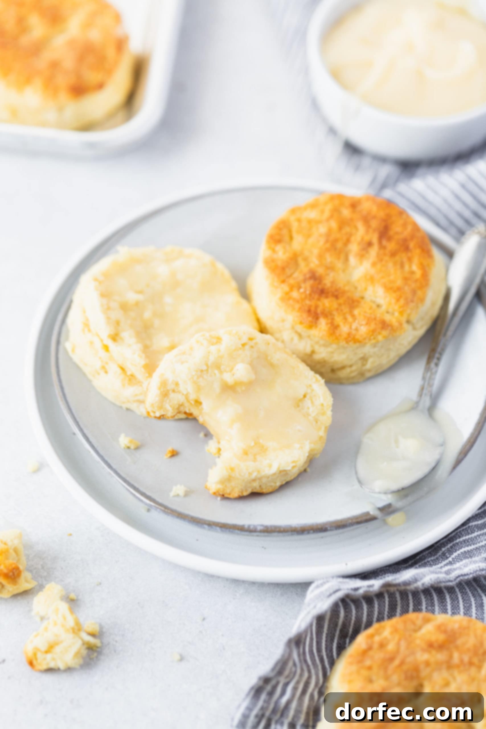 Two buttermilk biscuits on a white plate. One biscuit is cut in half and spread with honey butter. A bite has been taken out of one biscuit half.