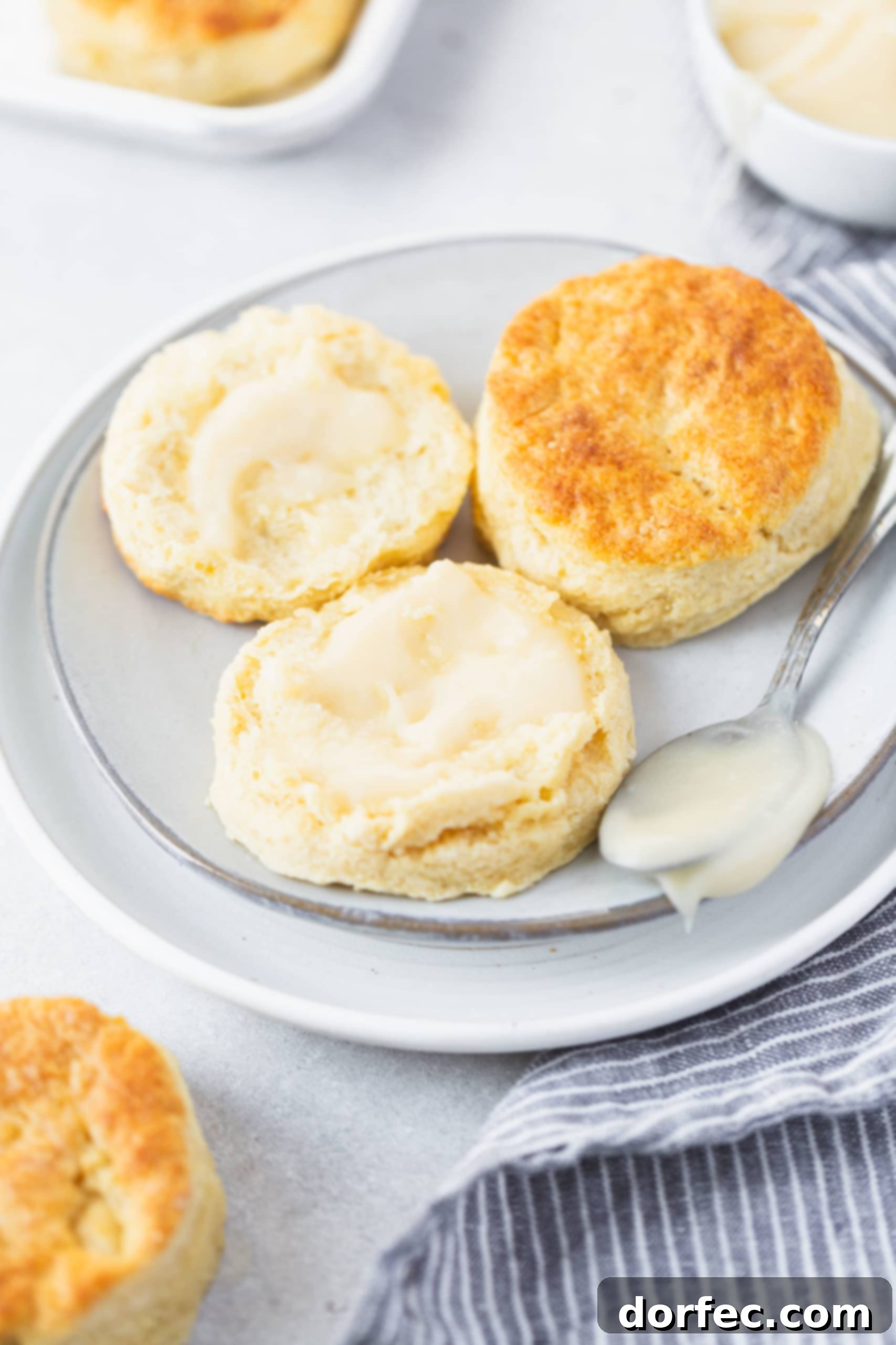 Two buttermilk biscuits on a small white plate. One biscuit is broken in half and has been buttered.