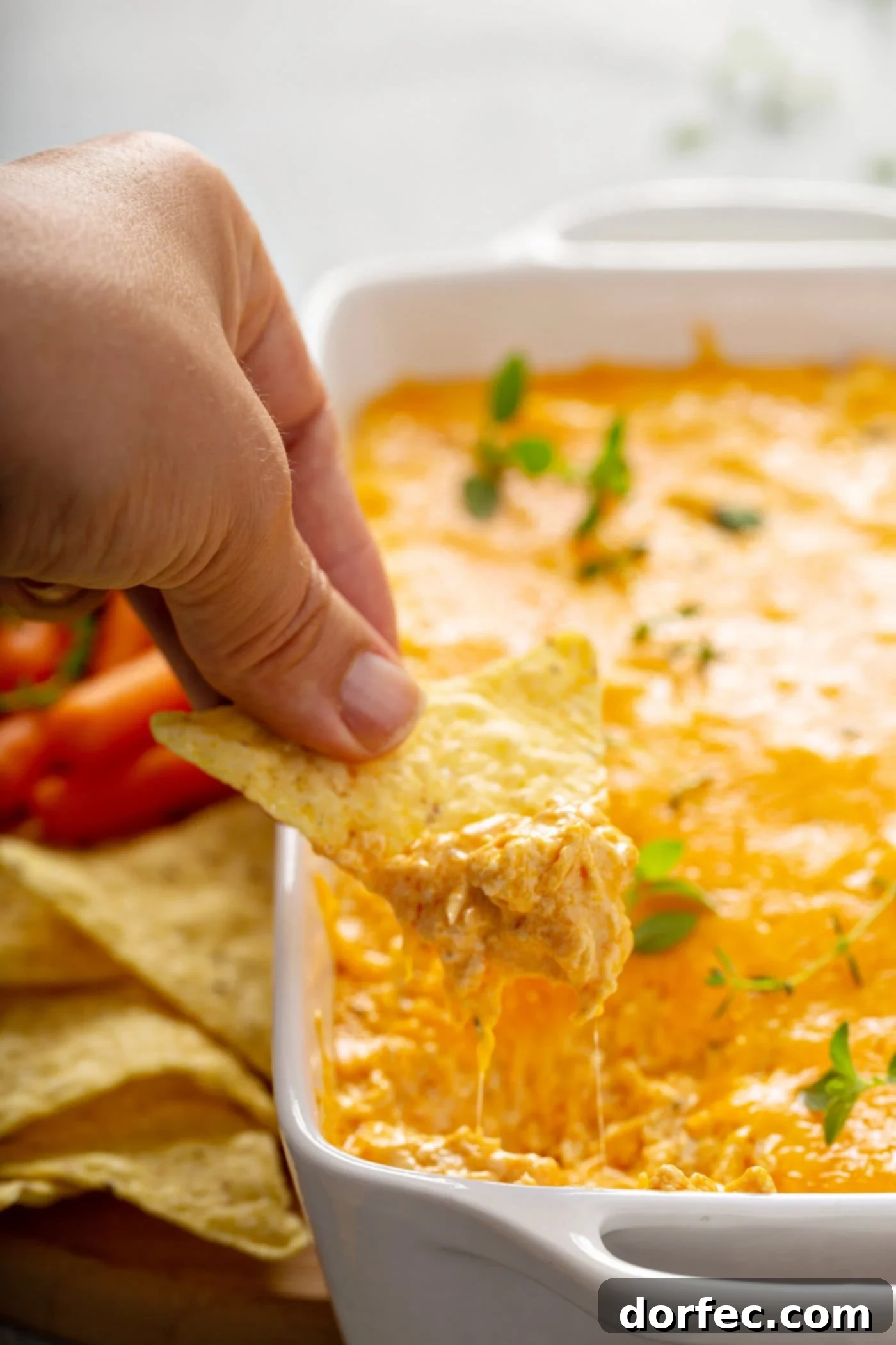 A crisp tortilla chip is being dipped into a warm, gooey Instant Pot Buffalo Chicken Dip. In the background, there are other chips and fresh carrot sticks, ready for dipping.