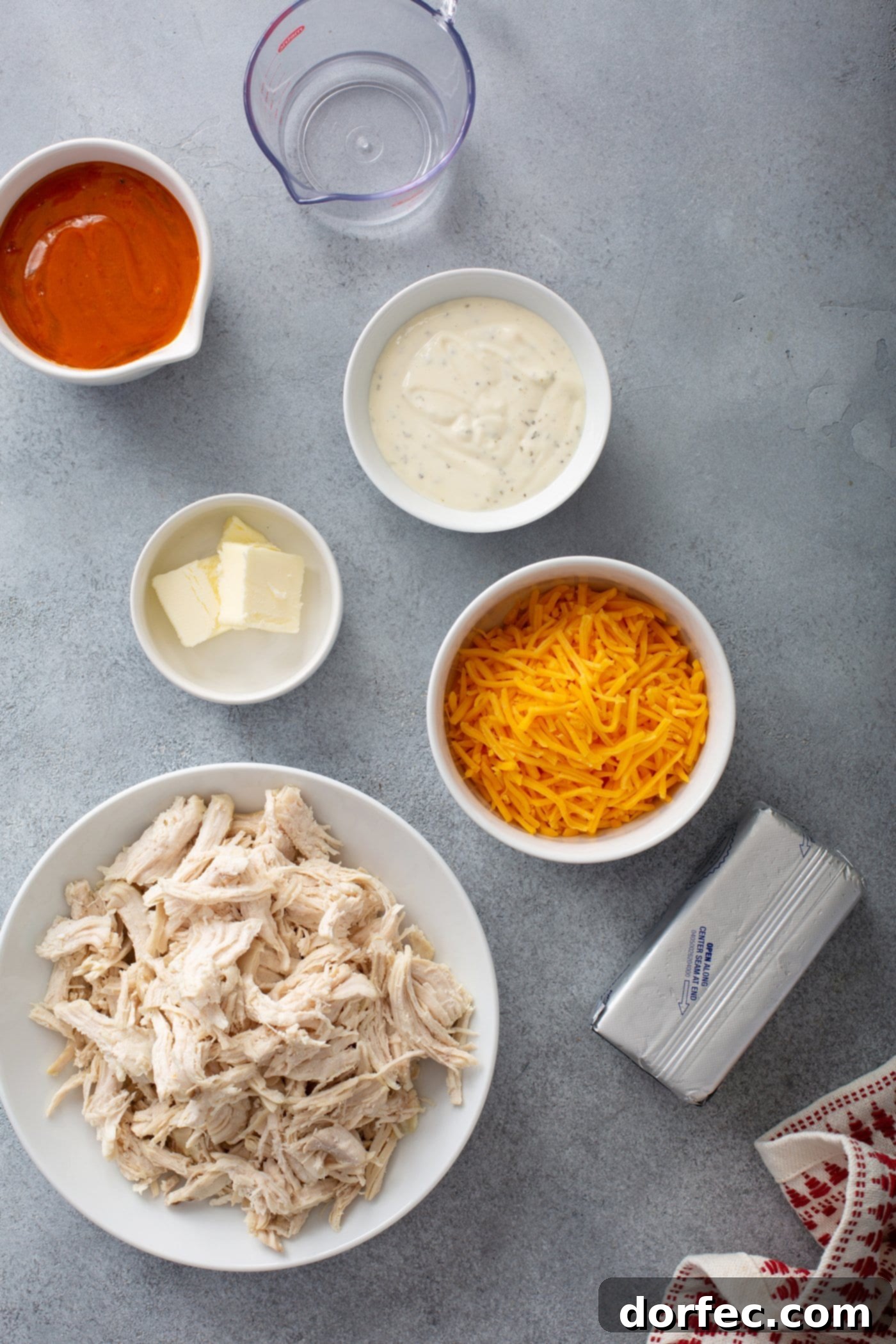 Small containers holding the key ingredients for Instant Pot Buffalo Chicken Dip: shredded chicken, a bowl of shredded cheese, a bottle of hot sauce, a bowl of ranch dressing, and a stick of butter.