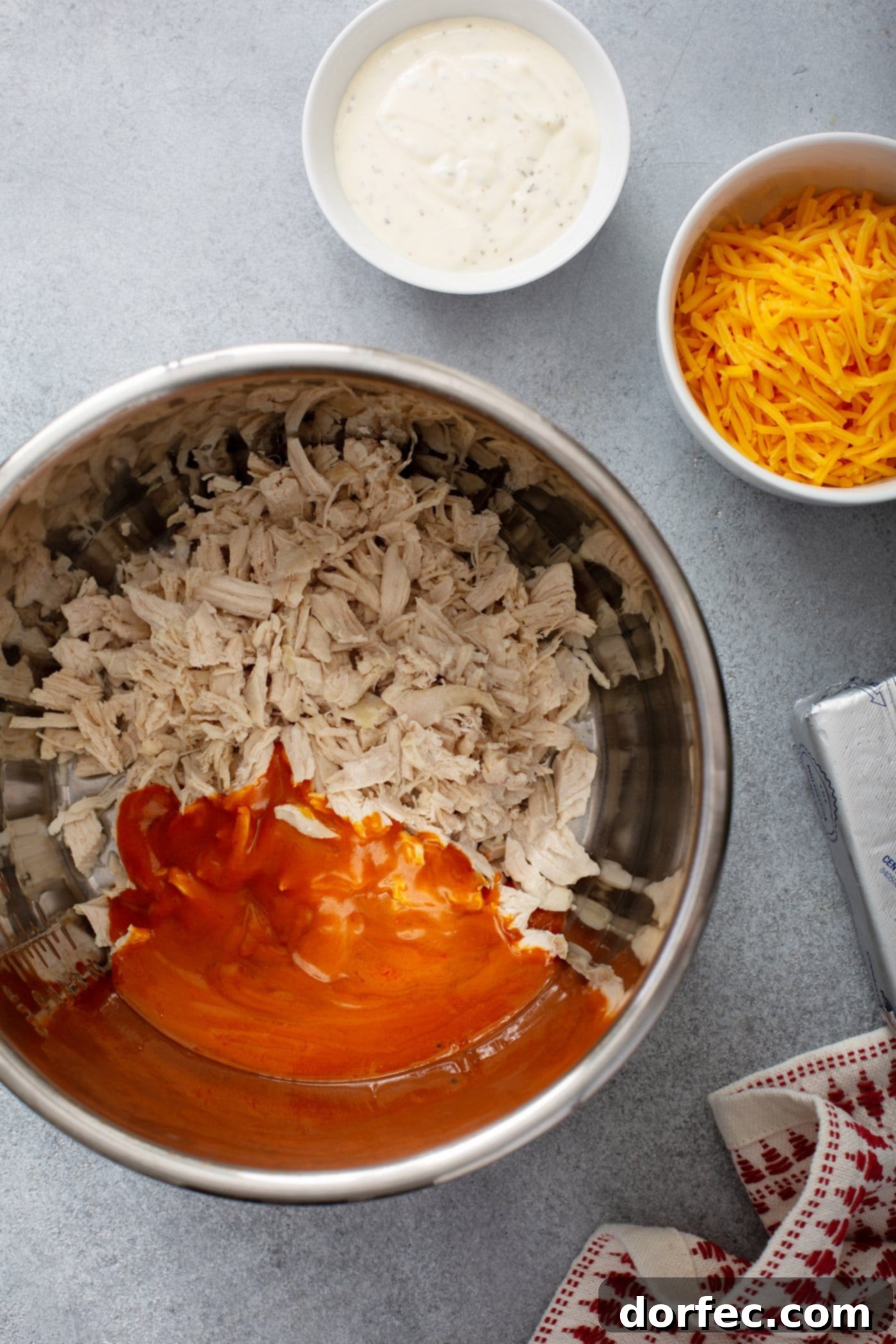 Inside an Instant Pot, cooked shredded chicken is being mixed with buffalo sauce. In the background, small bowls contain ranch dressing and shredded cheddar cheese, ready for the next step.