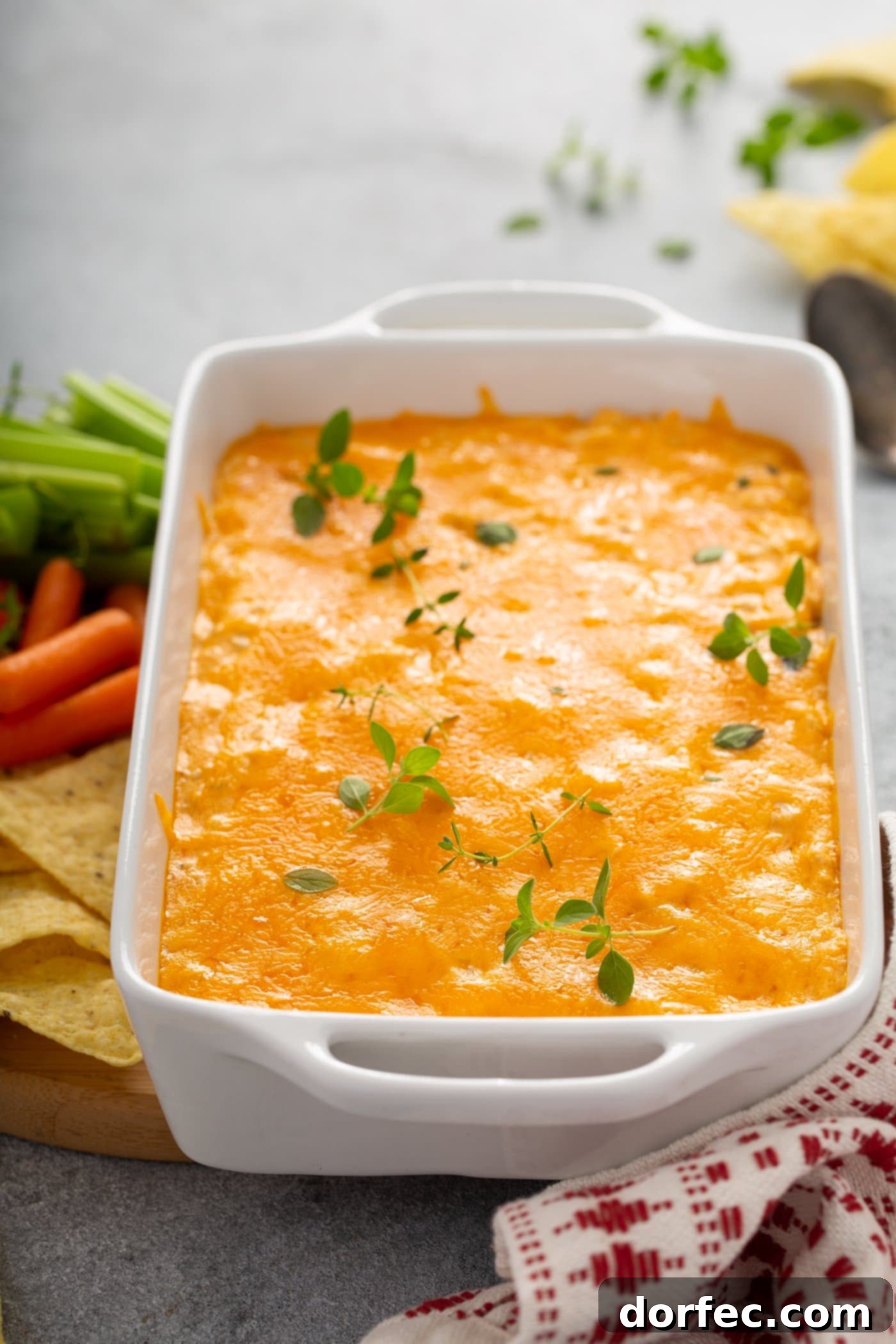 A bubbling casserole dish filled with hot Instant Pot Buffalo Chicken Dip, generously covered with melted cheese and sprinkled with fresh parsley. Beside the dish are crispy chips, fresh carrots, and crunchy celery for dipping.
