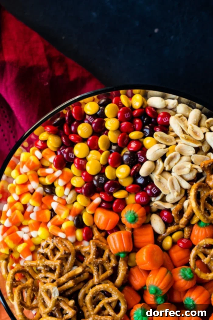 A beautifully arranged glass bowl brimming with a festive 5-Minute Halloween Snack Mix, featuring a colorful array of candy corn, salted peanuts, Halloween-themed M&Ms, crunchy pretzels, and charming candy pumpkins, set against a blurred background.