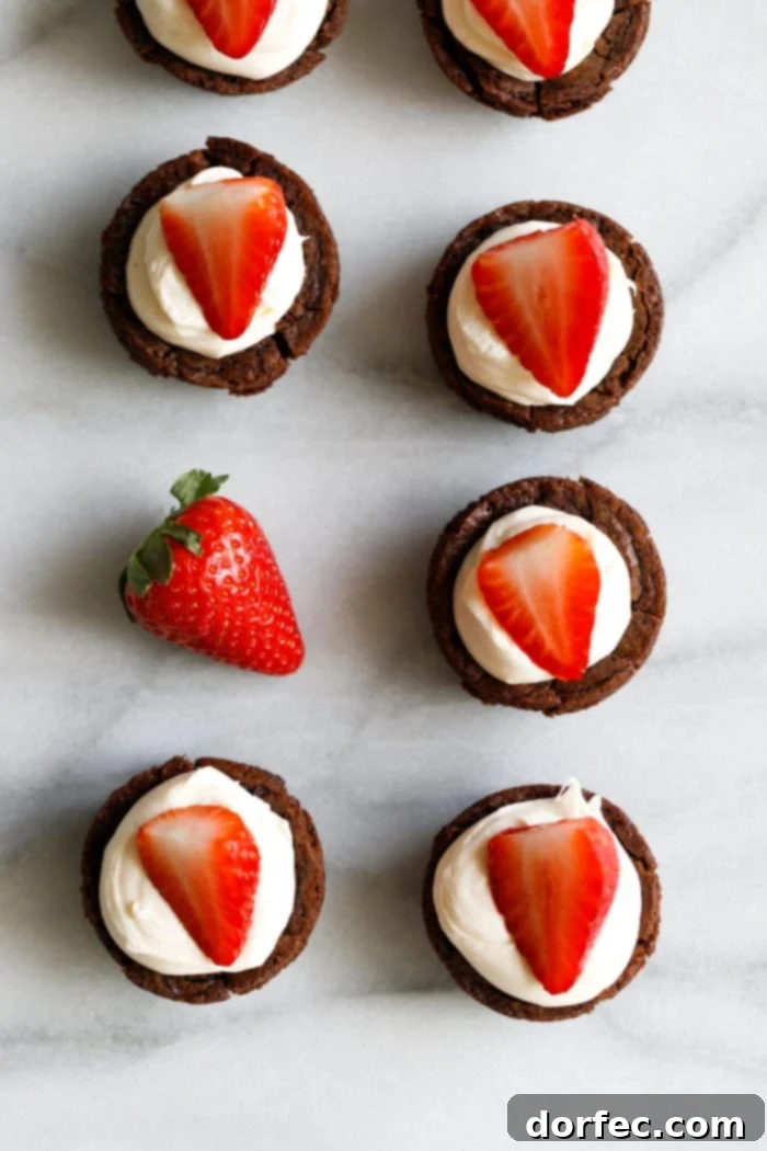 A marble background with tiny brownie cups filled with a white chocolate cream cheese filling and a sliced strawberry on top making these the cutest tuxedo brownie cups