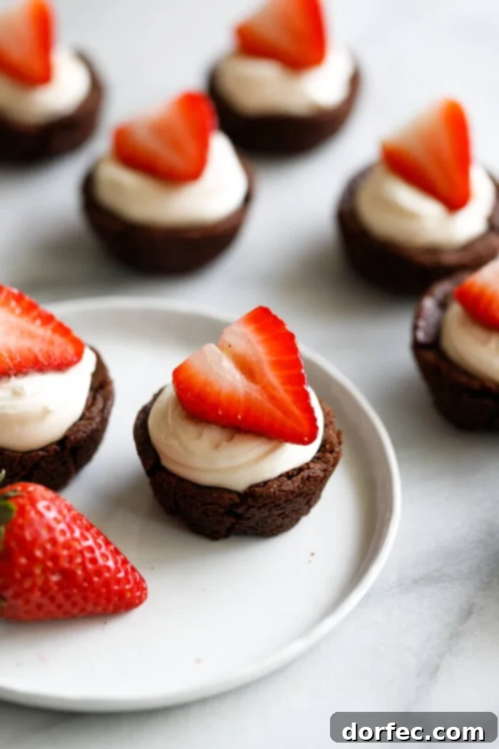 A marble background with a white plate filled with tiny brownie cups filled with a white chocolate cream cheese filling and a sliced strawberry on top making these the cutest tuxedo brownie cups