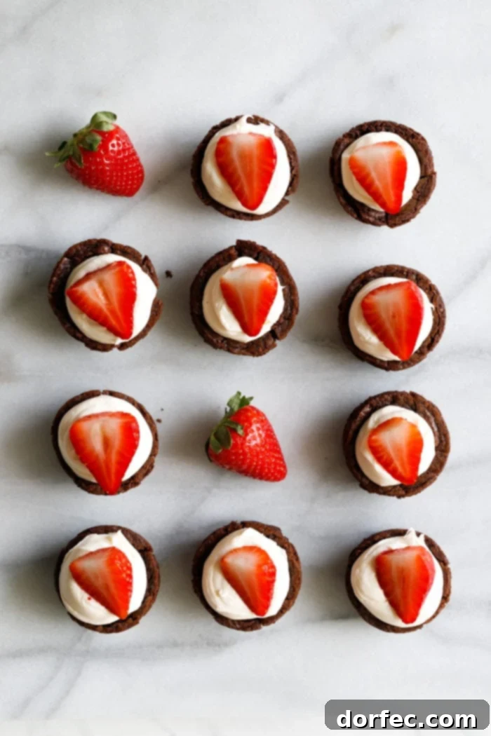 A marble background with a white plate filled with tiny brownie cups filled with a white chocolate cream cheese filling and a sliced strawberry on top making these the cutest tuxedo brownie cups