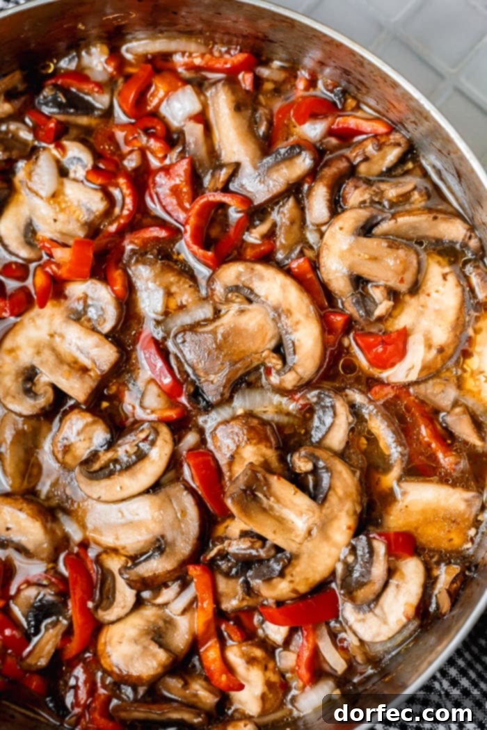 A photo of saucy sauteed baby bella mushrooms with sliced red bell peppers and onions in a saute pan, ready to be served.