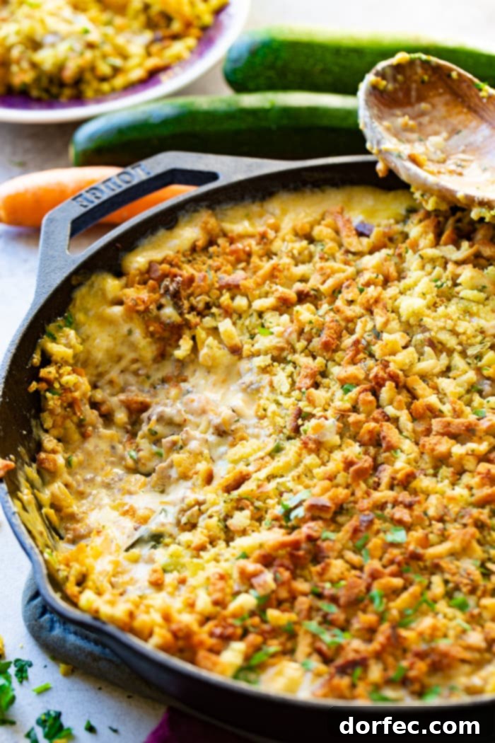 A close-up view of a creamy zucchini sausage casserole, topped with stuffing, in a cast iron skillet, with a scoop removed. The rich texture of the casserole and the crispness of the topping are visible.