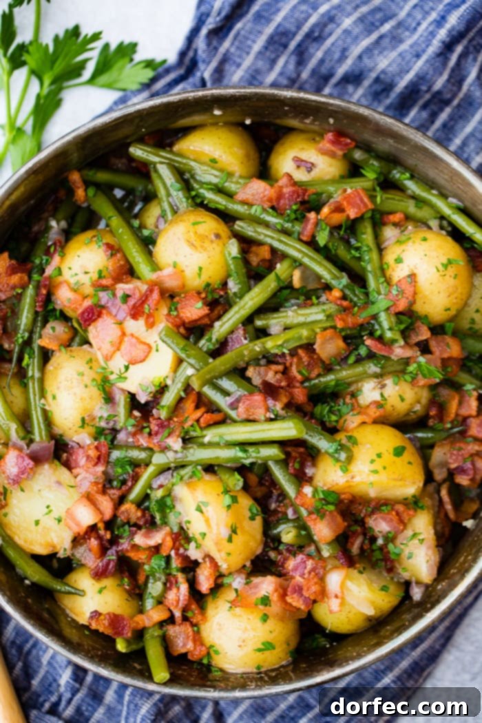 A silver serving dish filled with cooked fresh green beans, potatoes, and crispy bacon pieces, garnished with fresh parsley, sitting on a blue cloth napkin.