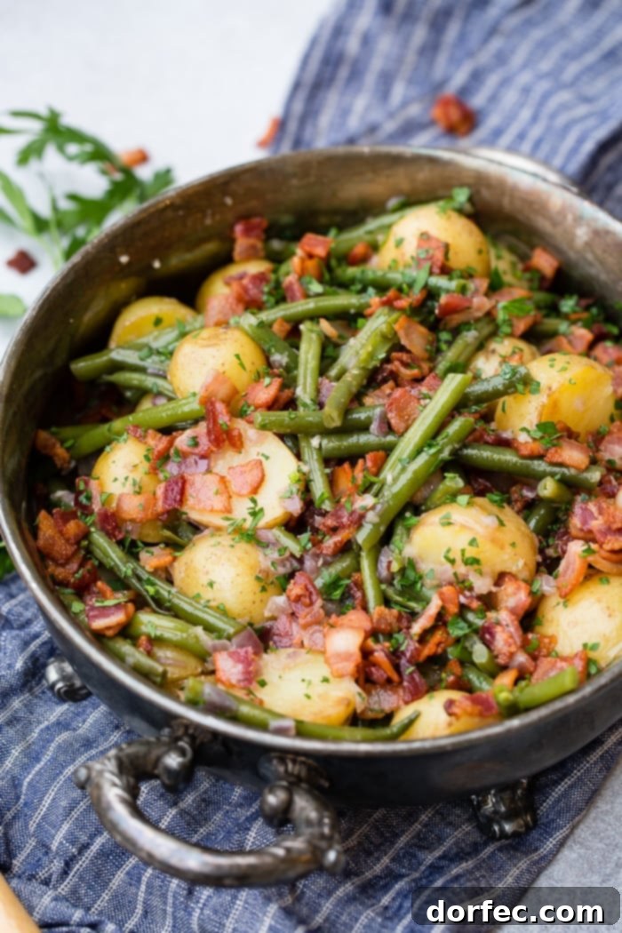 A photo of a silver serving dish sitting on a blue cloth napkin that is full of cooked fresh green beans, potatoes, and bacon pieces sprinkled with fresh parsley.