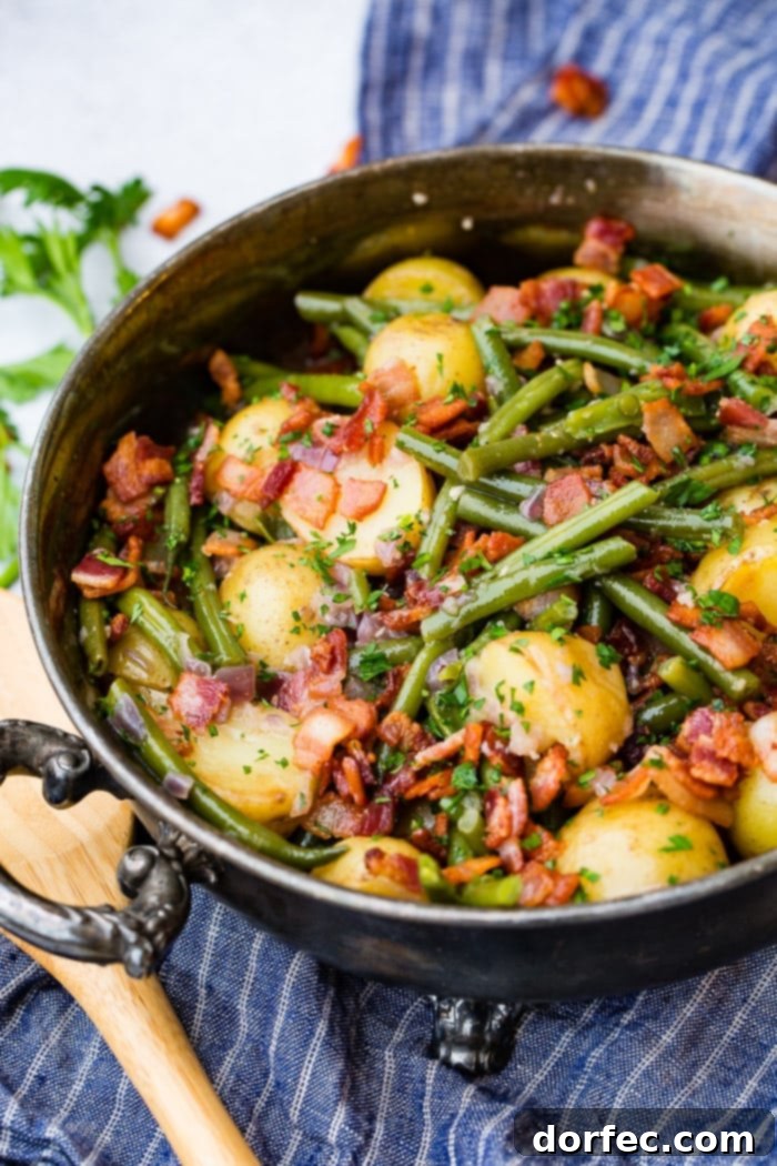 A photo of a large silver serving dish full of cooked green beans, halved potatoes, and crispy pieces of bacon, garnished with chopped parsley.