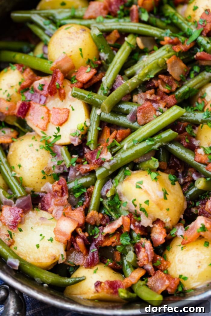 A zoomed-in photo of cooked green beans, potatoes, and bacon pieces sprinkled with fresh parsley, in a silver serving dish.