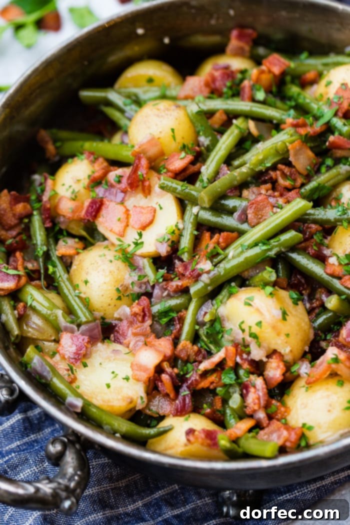 A photo of a silver serving dish sitting on a blue cloth napkin that is full of cooked fresh green beans, potatoes, and bacon pieces sprinkled with fresh parsley.