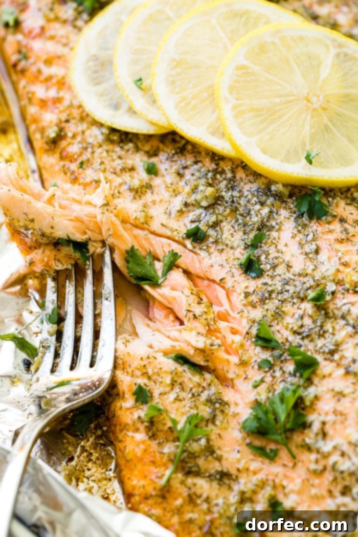 A close-up shot of a baked salmon fillet, flaked perfectly with a fork, garnished with fresh dill and parsley, sitting on aluminum foil