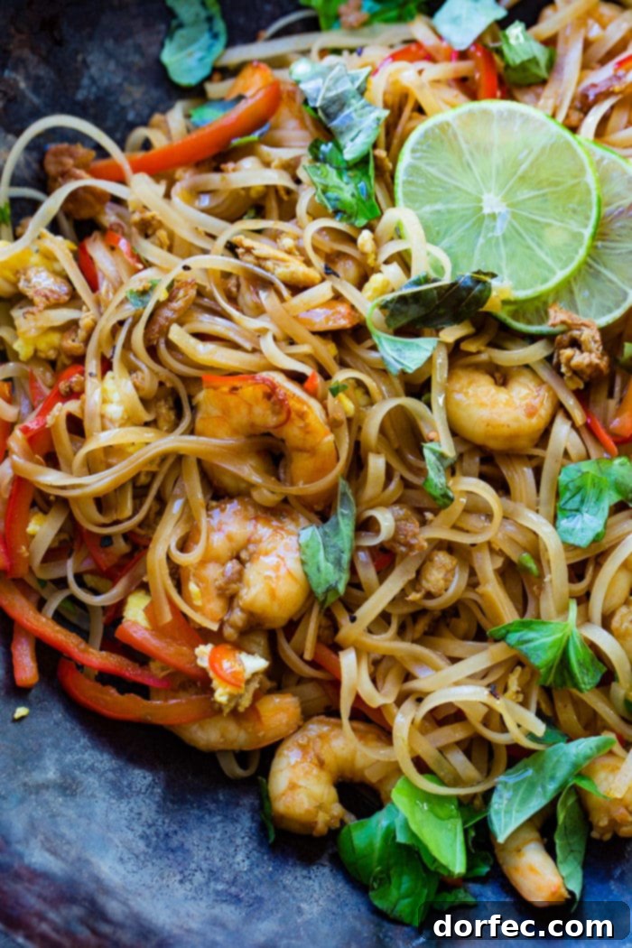 A photo of cooked shrimp, rice noodles, red bell peppers, and fresh basil leaves on a blue plate garnished with slices of lime.