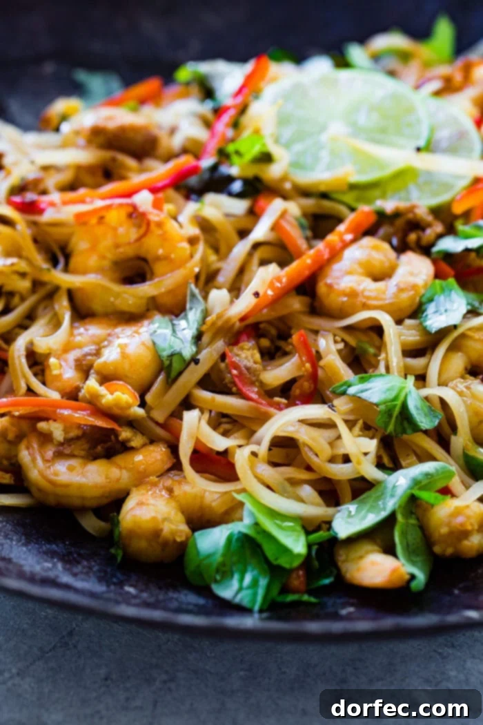 A photo of cooked shrimp, rice noodles, red bell peppers, and fresh basil leaves on a blue plate garnished with slices of lime.