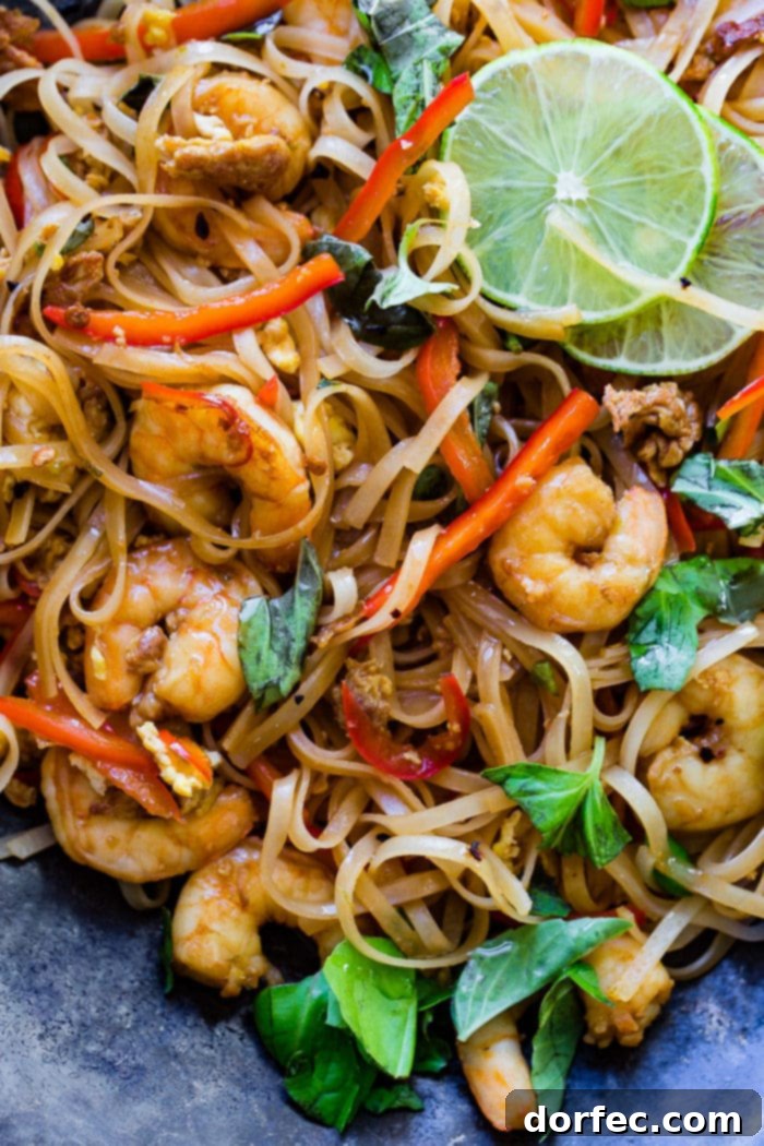 A photo of cooked shrimp, rice noodles, red bell peppers, and fresh basil leaves on a blue plate garnished with slices of lime.